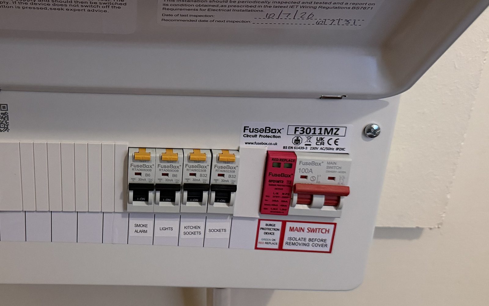 Consumer unit with the lid lifted, showing a row of labelled circuit breakers and, on the right, the red main switch marked Isolate before removing cover