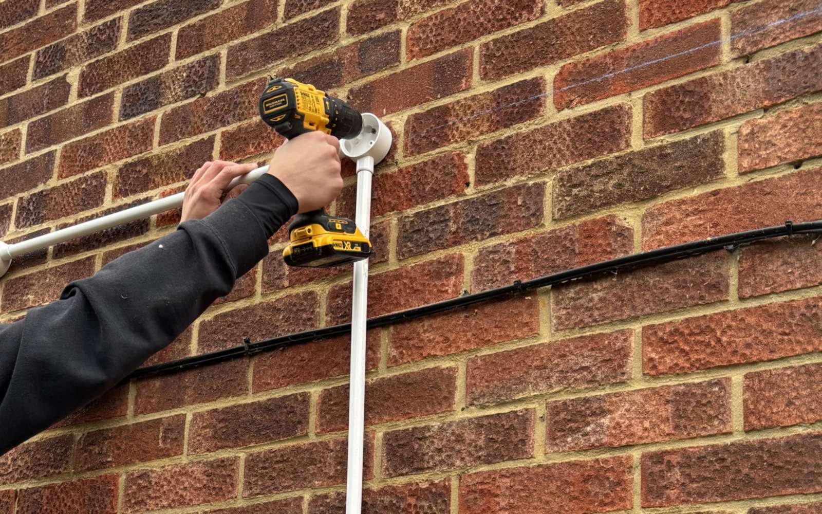 White PVC conduit box being fixed to the outside brick wall of a house with a cordless drill, the conduit run dropping down the wall below