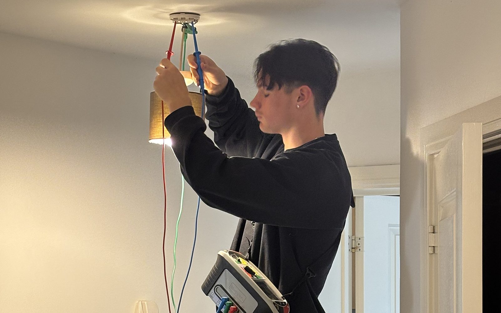 Trainee electrician Taylor Boon testing at a ceiling light, with test leads running down to a multifunction installation tester